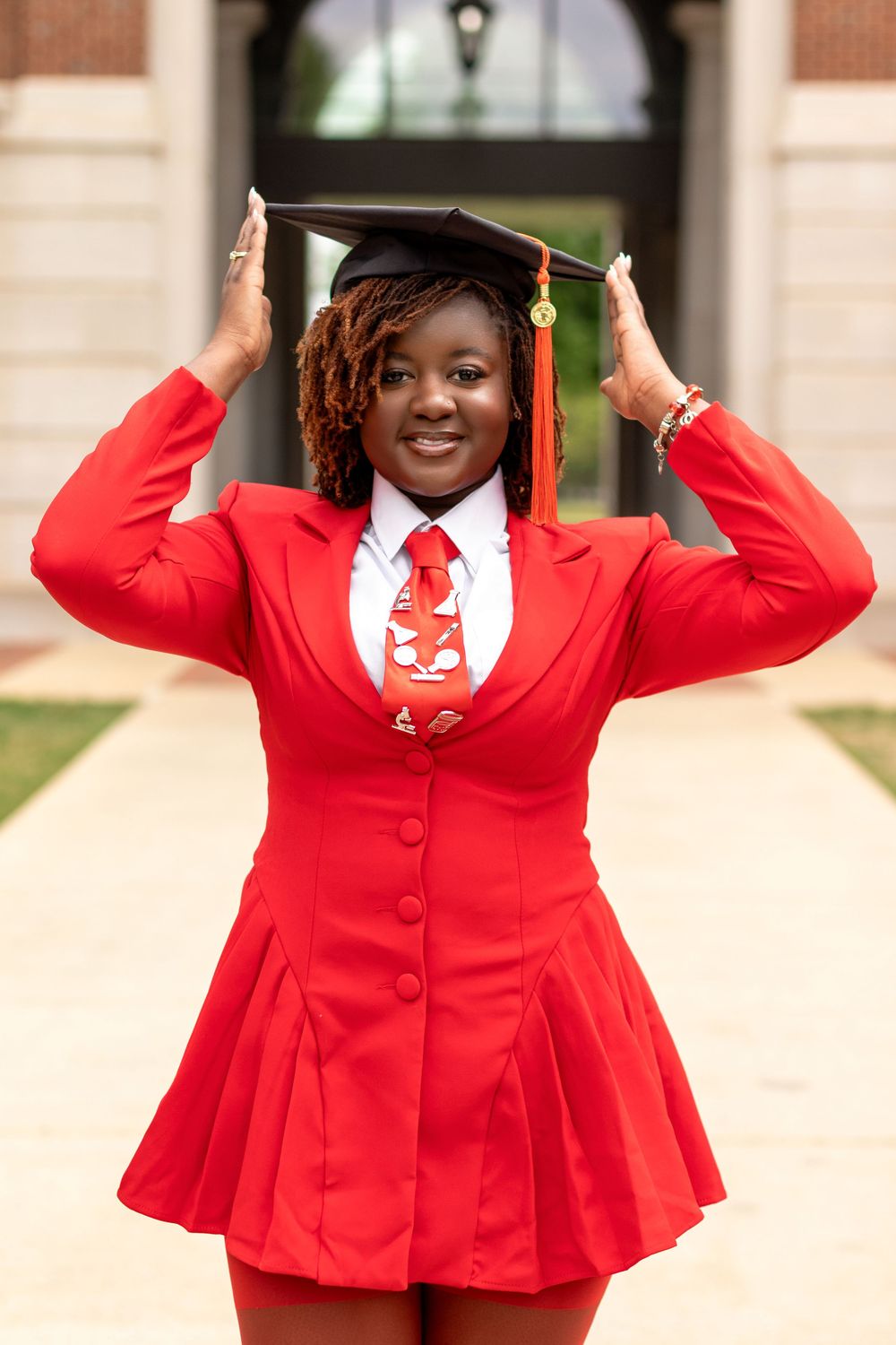 Smiling graduate adjusts black cap wearing red blazer dress with white shirt and red tie outdoors.