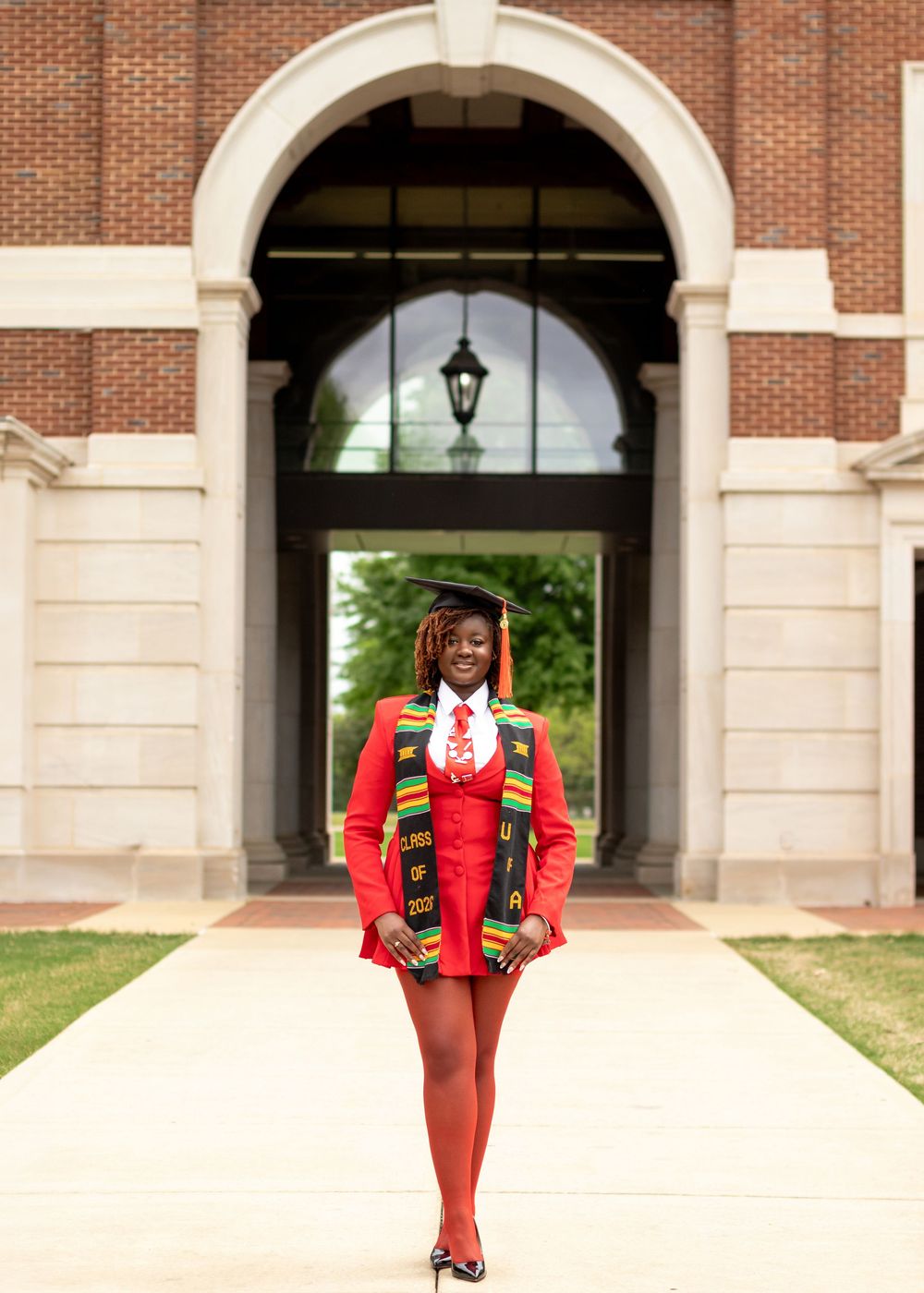 Graduate in red blazer dress and kente stole poses proudly under brick archway on college campus.