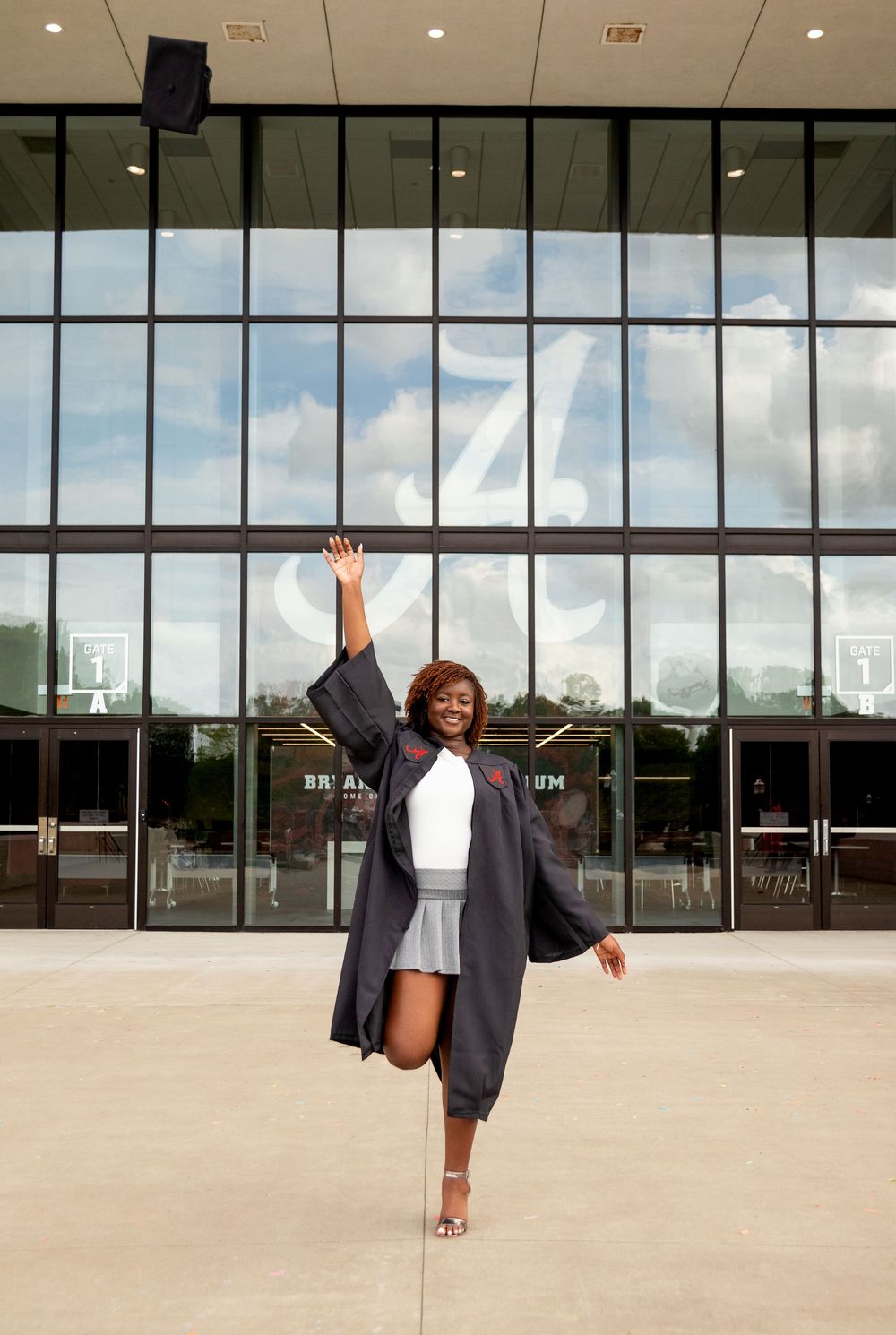 Joyful graduate in cap and gown tossing mortarboard in air outside modern university building.