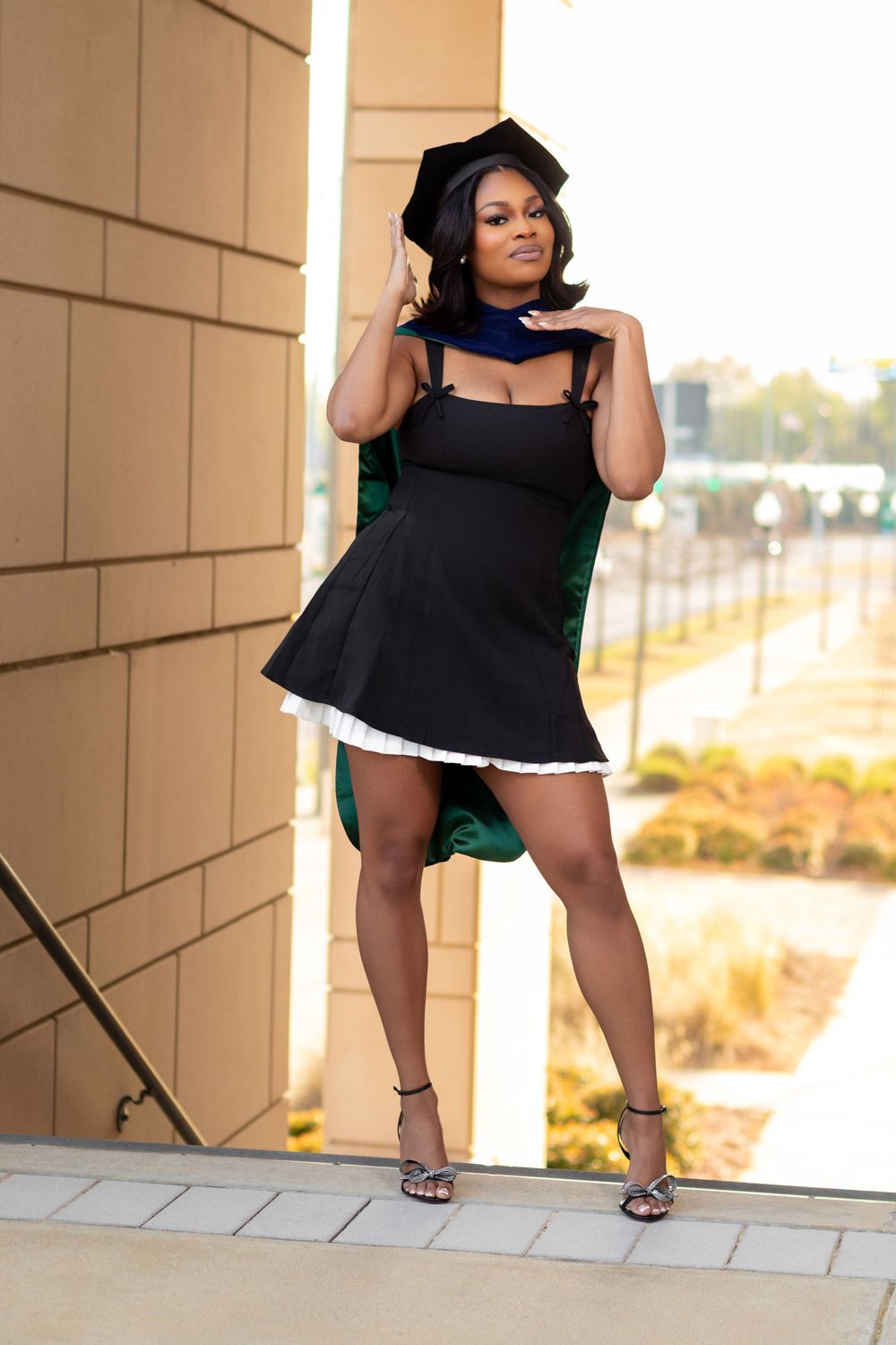 A young Black woman in a short black dress and graduation cap poses confidently outdoors near a building.