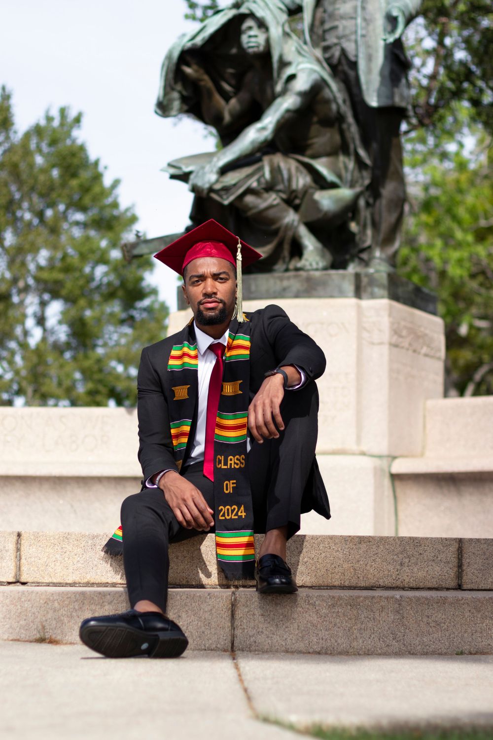 Graduate in traditional academic regalia poses near historic statue on campus steps.