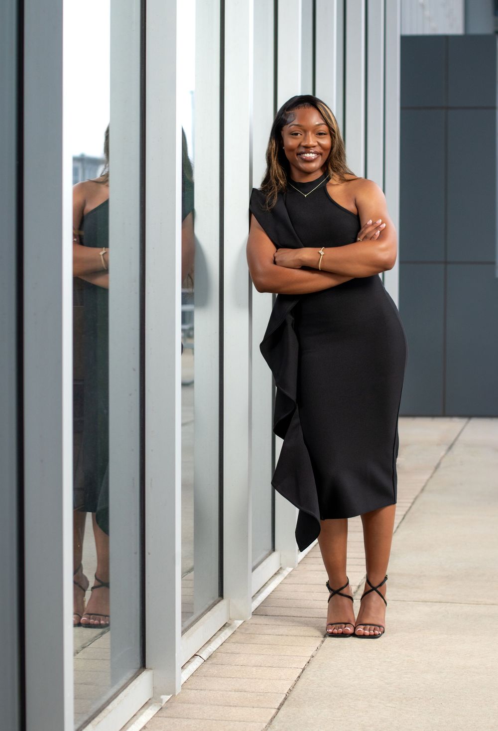 A confident woman in a black dress stands arms crossed against a modern glass building exterior.
