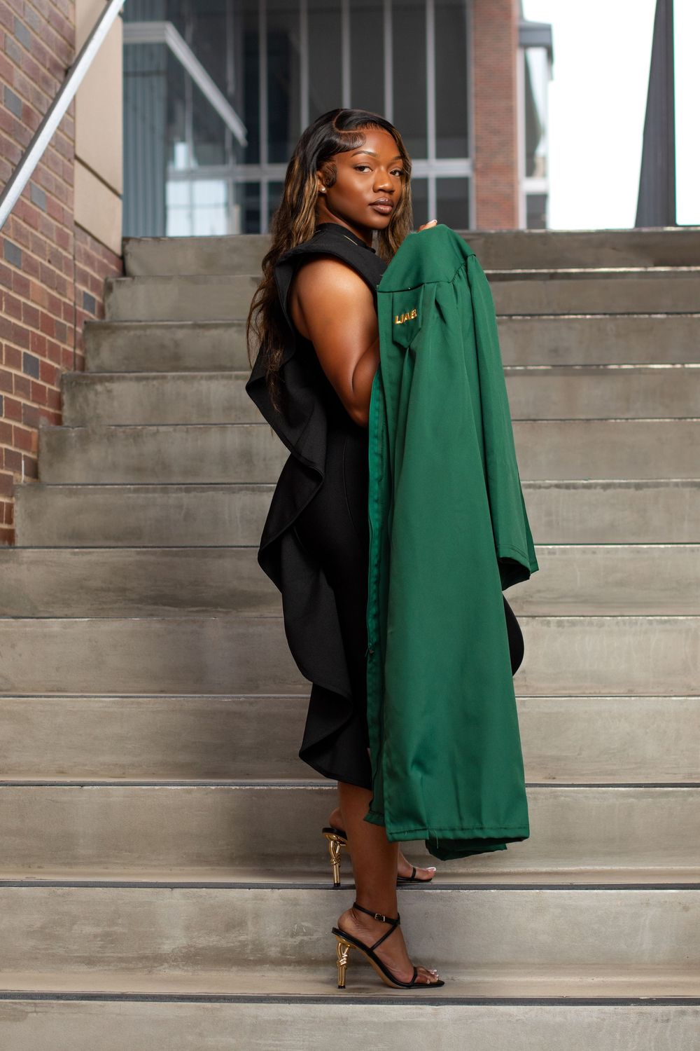 Young woman in black dress holding green graduation gown poses on stone steps outside a brick building.