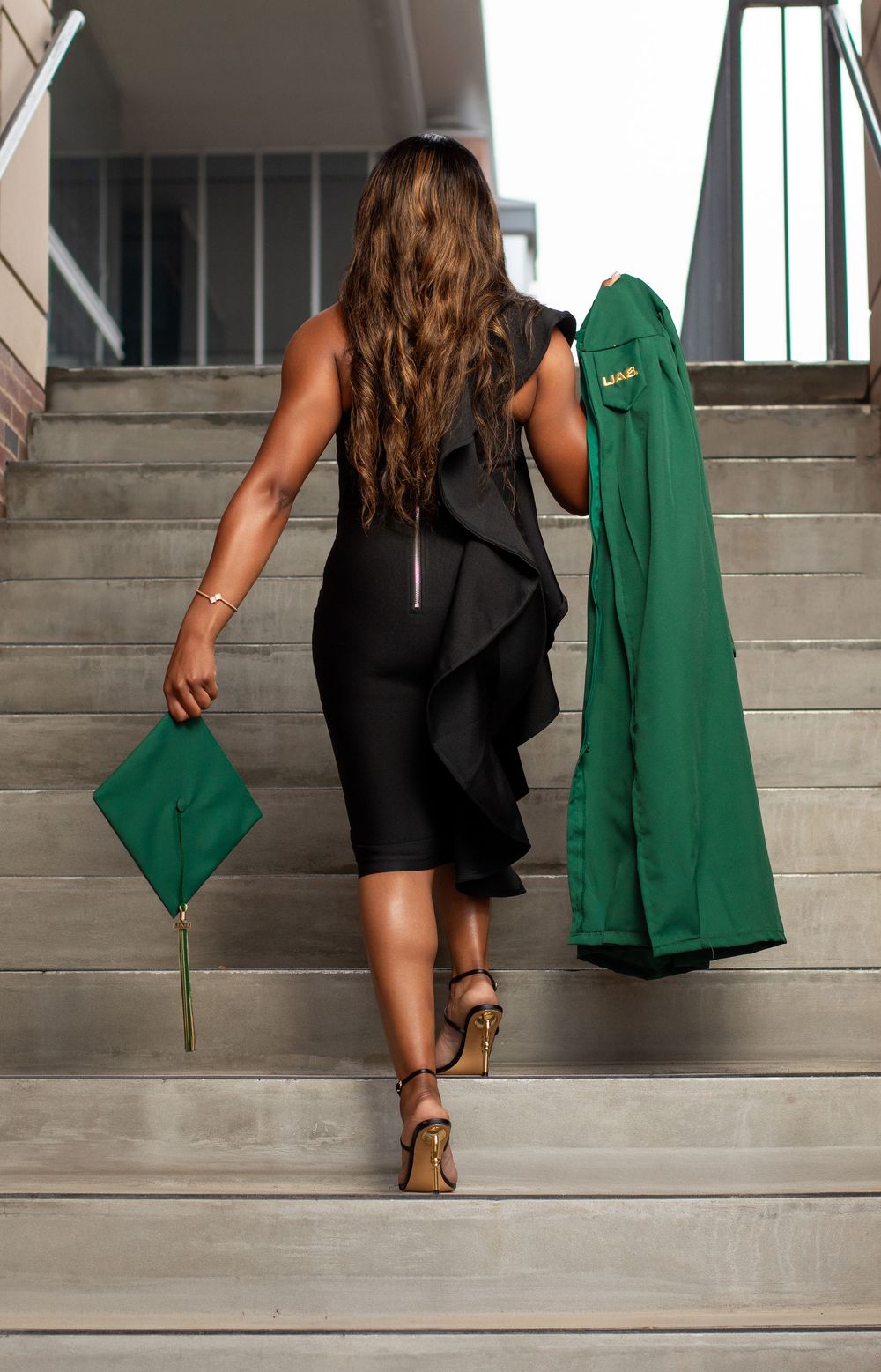 Woman in black dress carrying green graduation cap and gown walks up concrete stairs, viewed from behind.