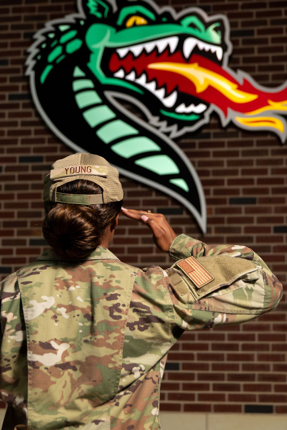 Military soldier in camouflage uniform saluting a dragon mascot mural on a brick wall.