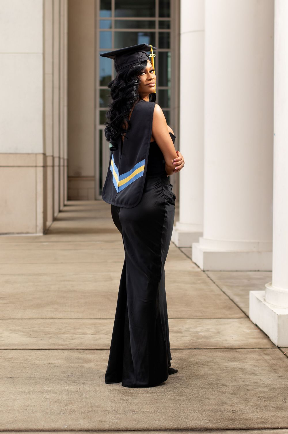 Young Black woman in graduation cap and gown poses elegantly in long black dress between white columns.