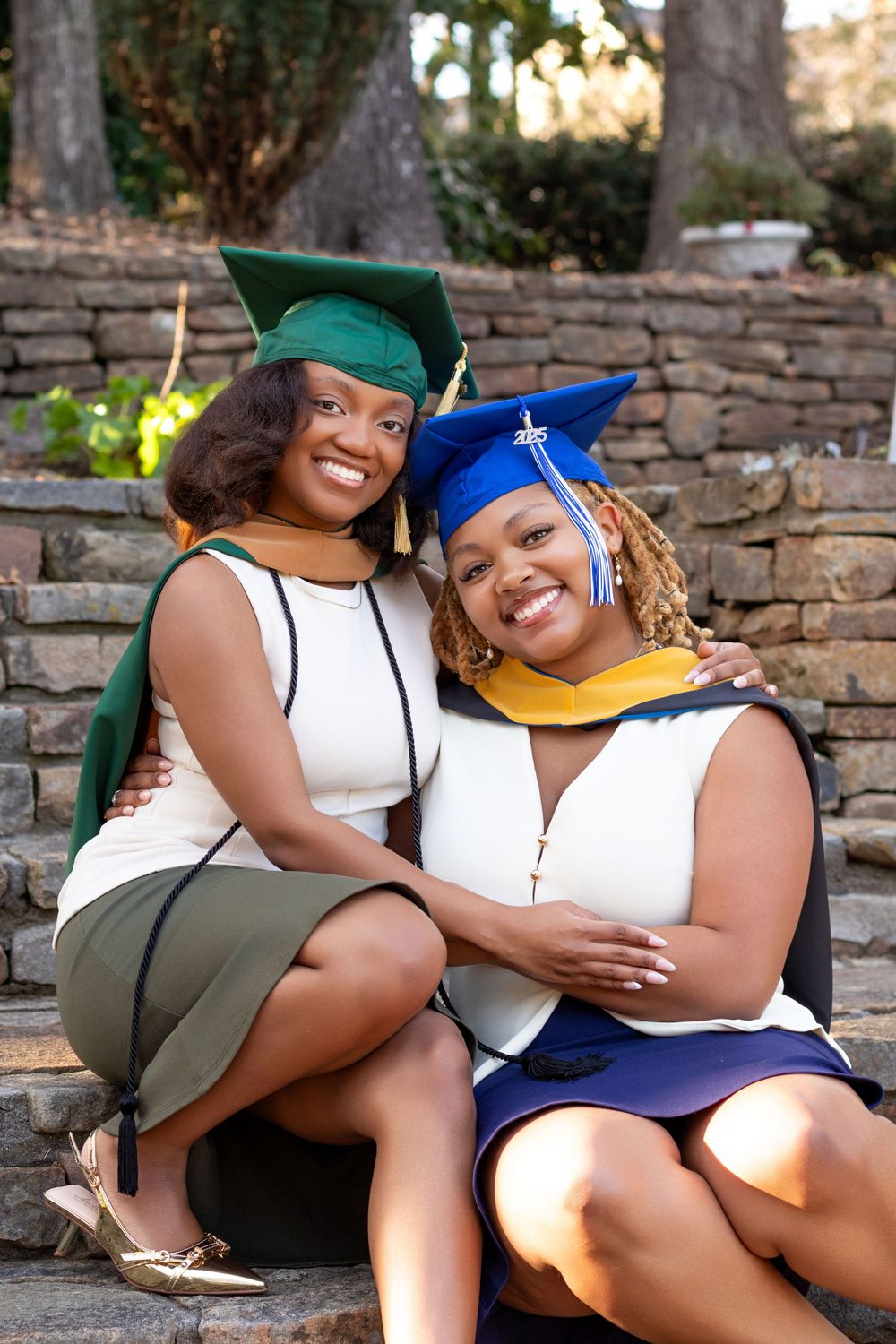 Two smiling women in graduation caps and gowns pose together on stone steps outdoors.