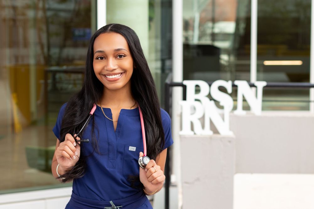 Smiling female nursing student in blue scrubs holding a stethoscope, standing near BSN RN sign outside university.
