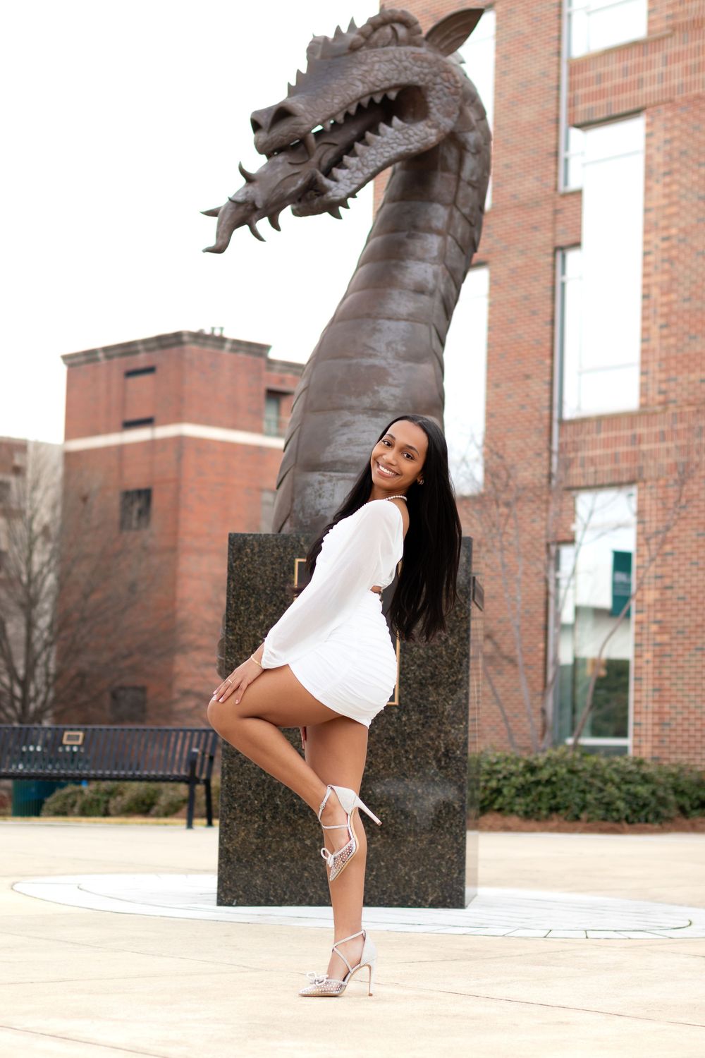 Woman in white mini dress posing confidently in front of a dragon sculpture on a college campus.