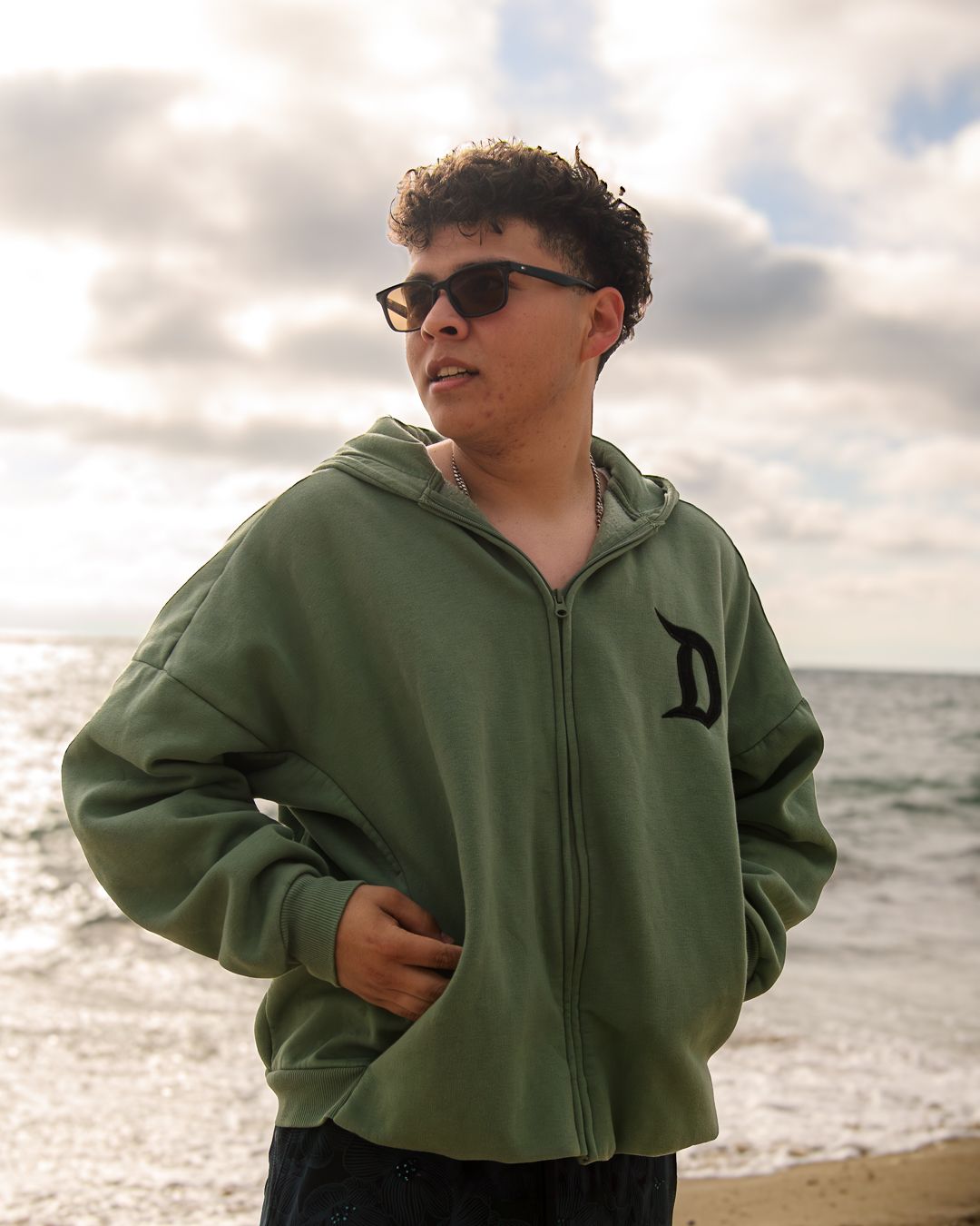 Young man in green zip-up hoodie with D logo wearing sunglasses at beach.