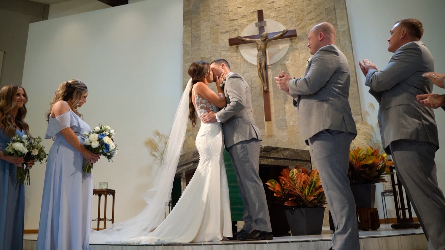 A couple exchanges vows at a church altar during a wedding ceremony surrounded by bridesmaids and groomsmen in grey.