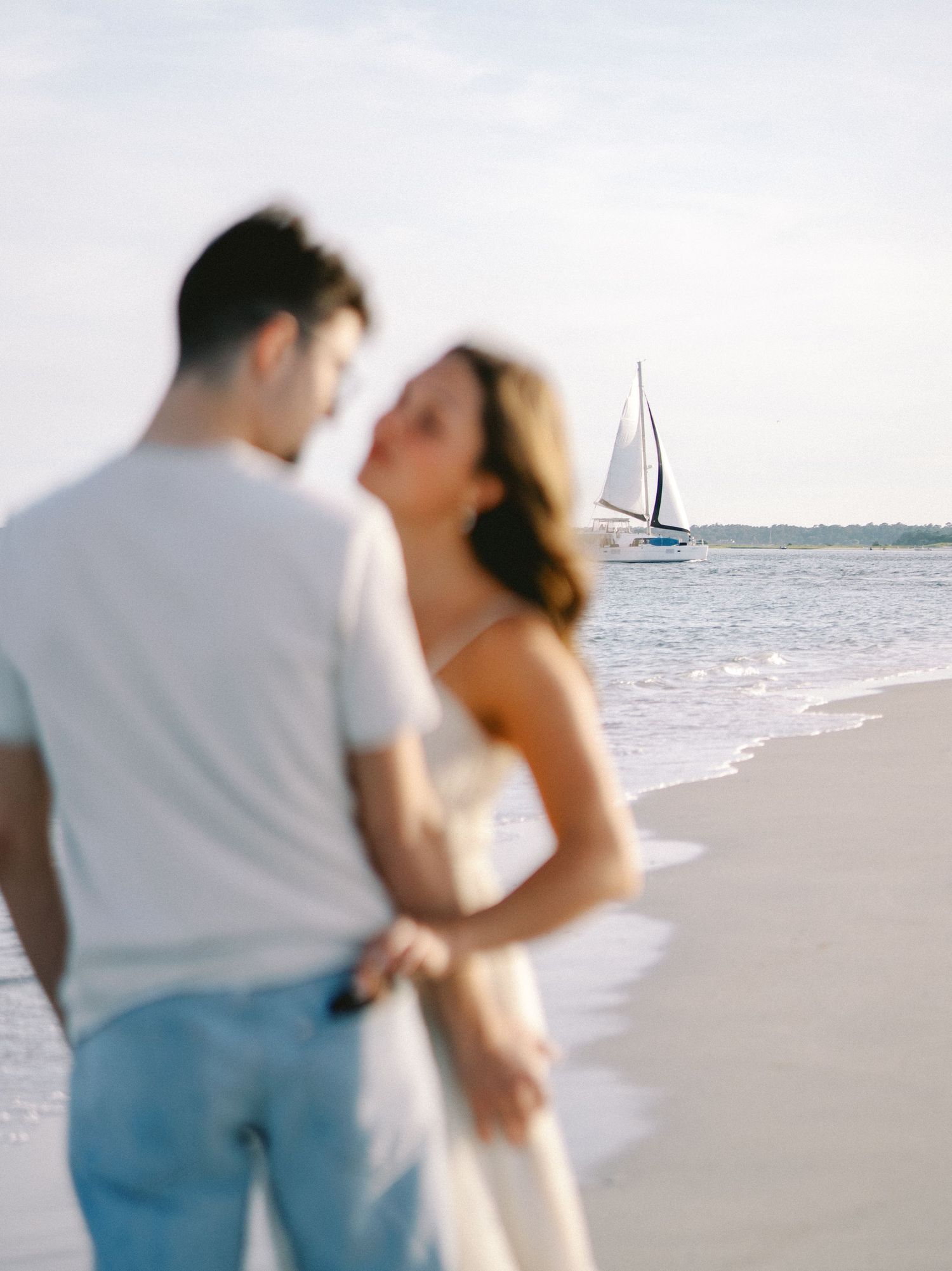 Couple in casual beach attire with sailboat visible on horizon.