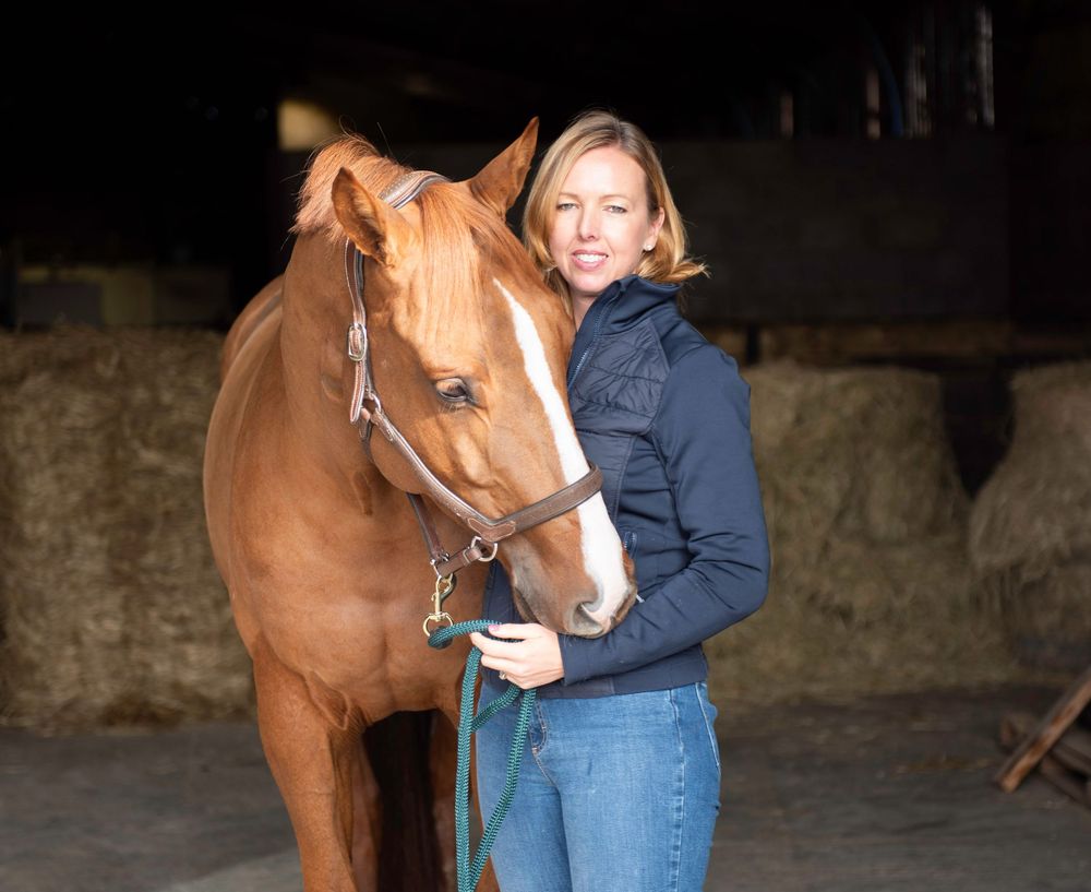 Brand photography - blonde woman wearing a dark blue jacket and jeans, standing next to her chestnut horse, and smiling