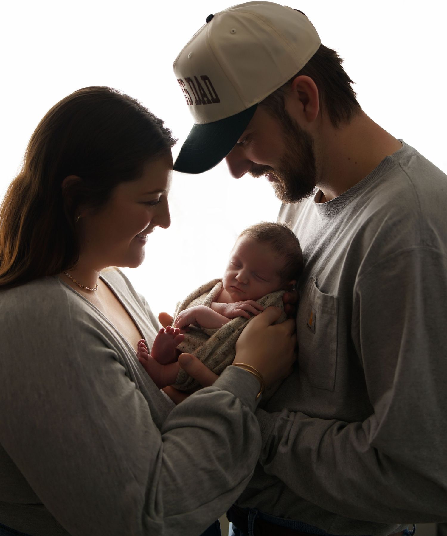 Silhouetted parents in gray shirts gazing lovingly at their sleeping newborn baby in a backlit newborn family portrait.