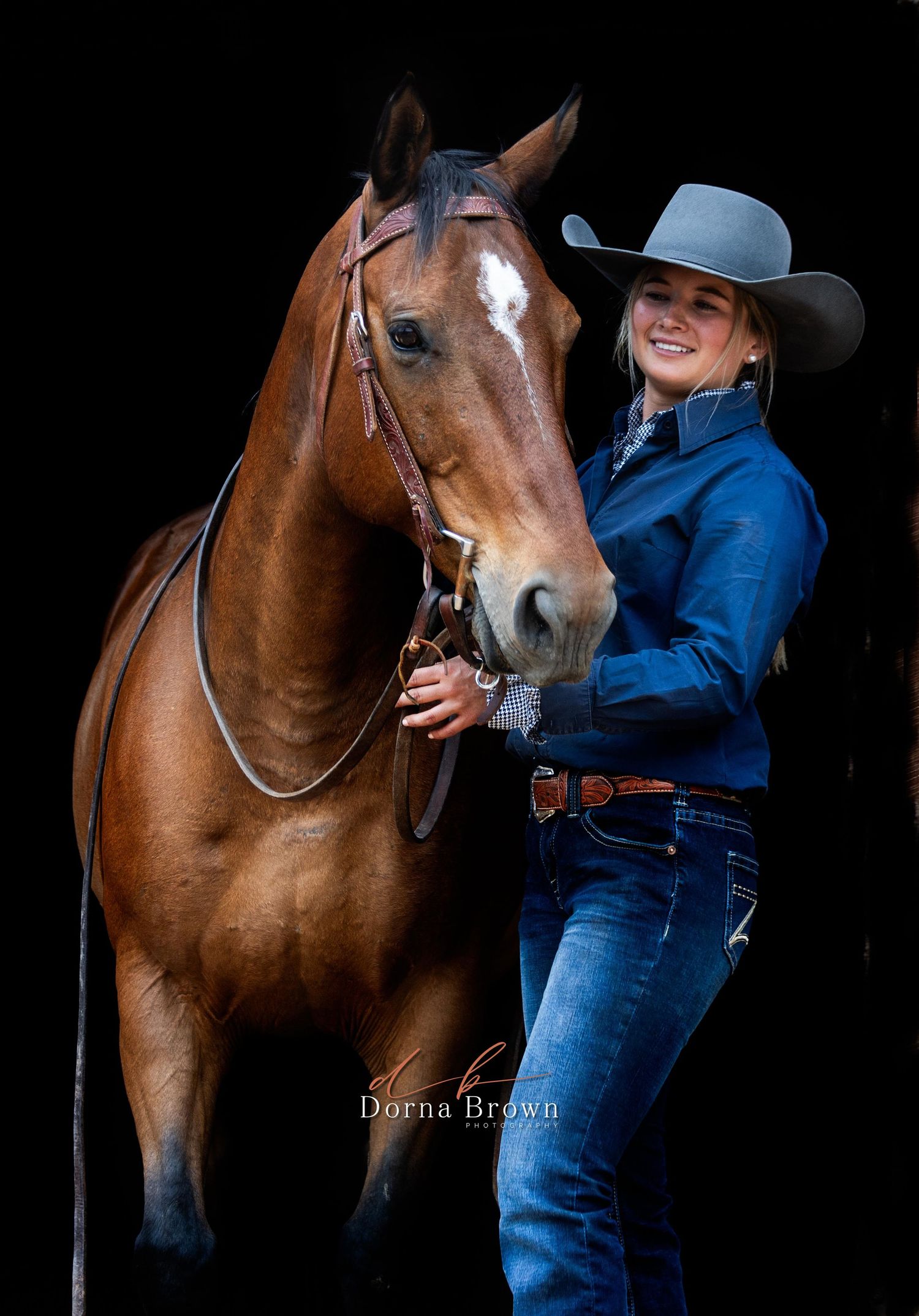 Western equestrian wearing blue denim and cowboy hat stands with beautiful chestnut horse against dark background.