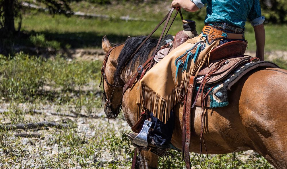 Close-up view of a western saddle with leather fringe and rider wearing turquoise shirt while horseback riding outdoors.