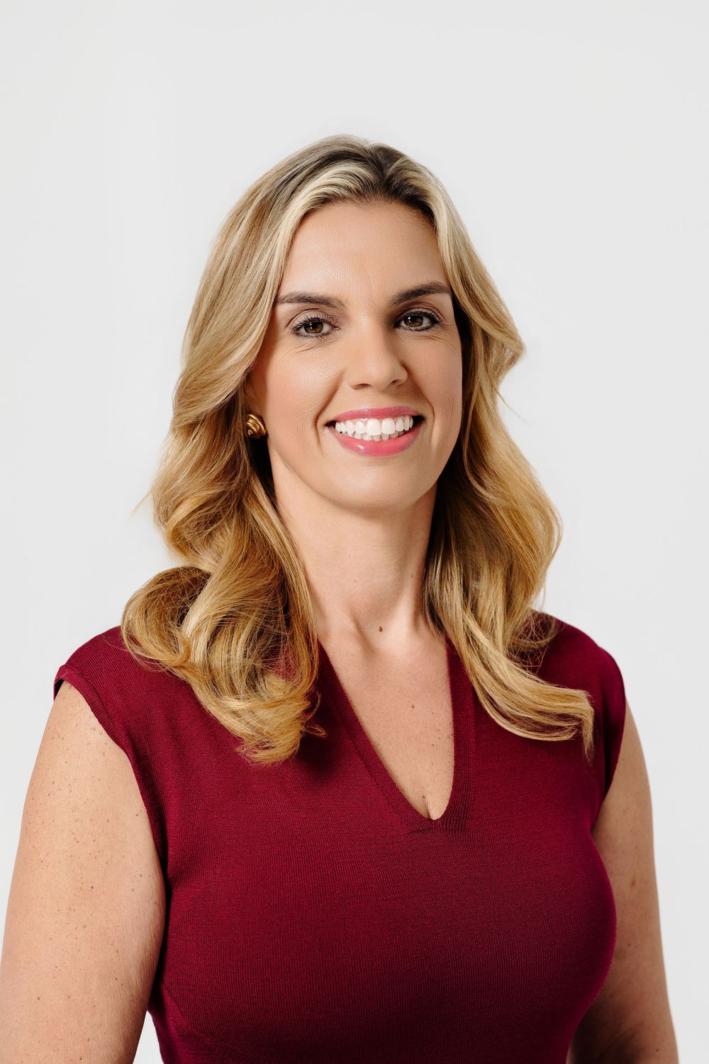 Smiling woman with long wavy blonde hair wearing a burgundy v-neck top and gold earrings in a professional studio portrait.