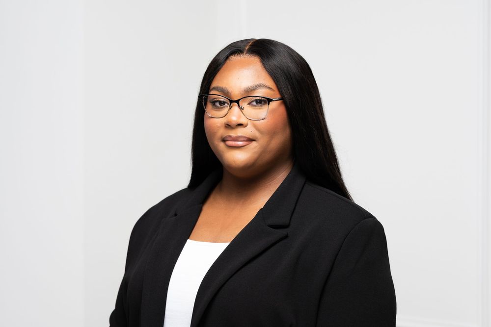 Smiling woman with long straight black hair and glasses wearing a black blazer over a white top against a light background.