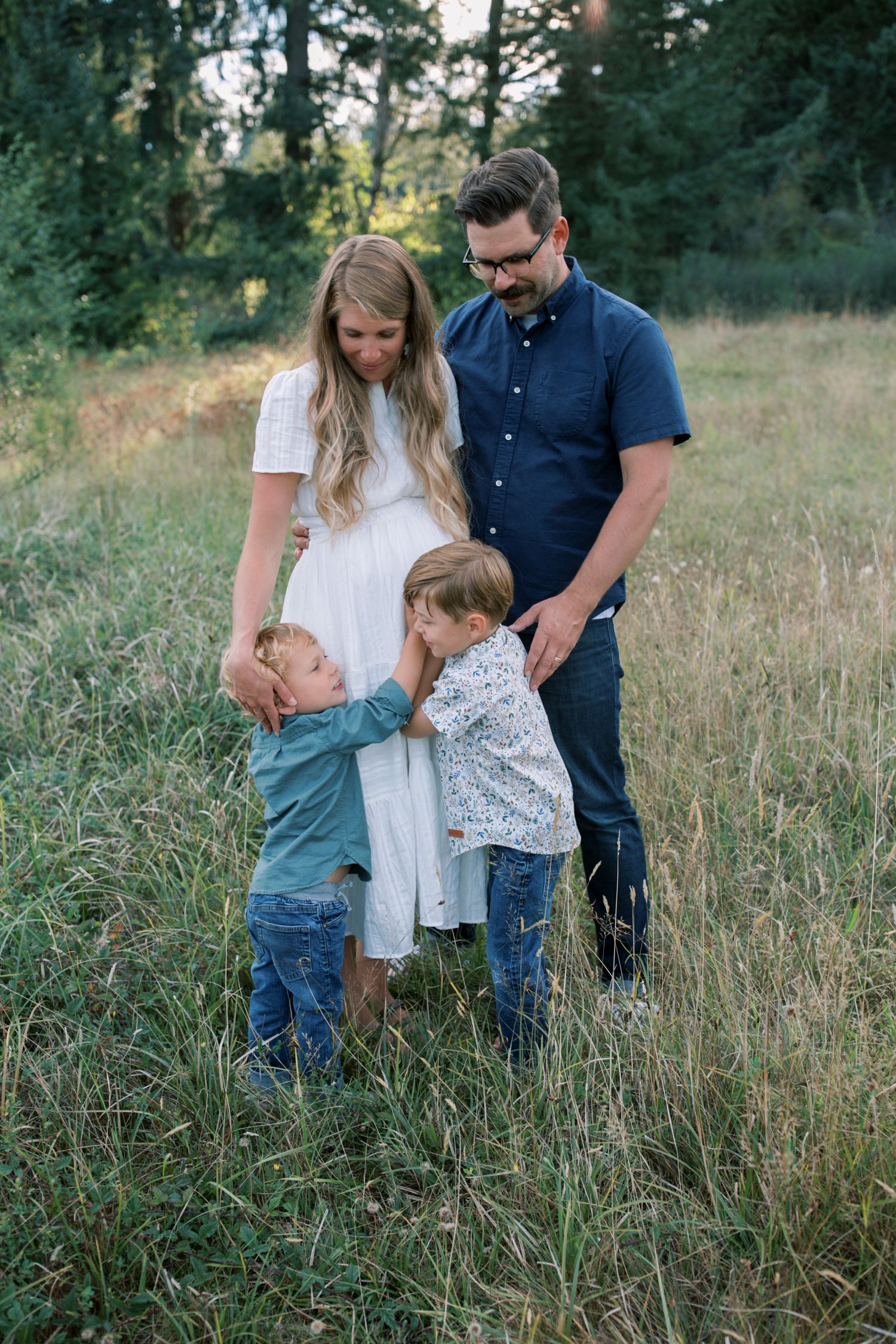 Family of four posing in a green meadow, parents watching two young boys interact playfully outdoors.