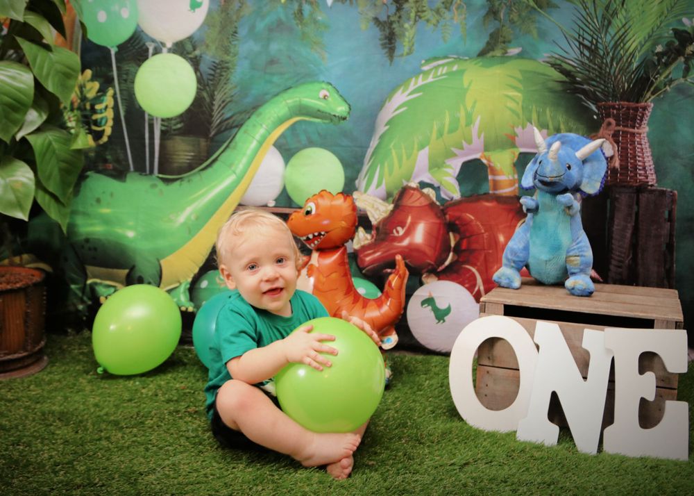 A dinosaur themed first birthday setup with green balloons, plush dinosaurs, and white ONE letters.