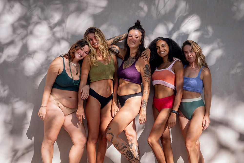 Group of diverse women in colorful swimwear pose together against textured shadow wall.