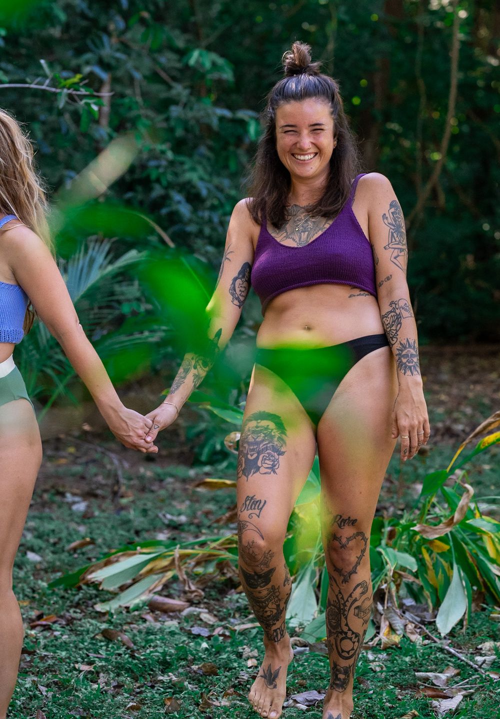 Person with colorful tattoos wearing purple top and green bottoms poses outdoors in natural setting.