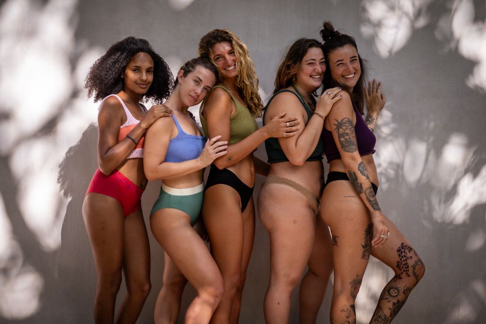 Group of friends in colorful swimwear posing together against white wall with dappled shadows.