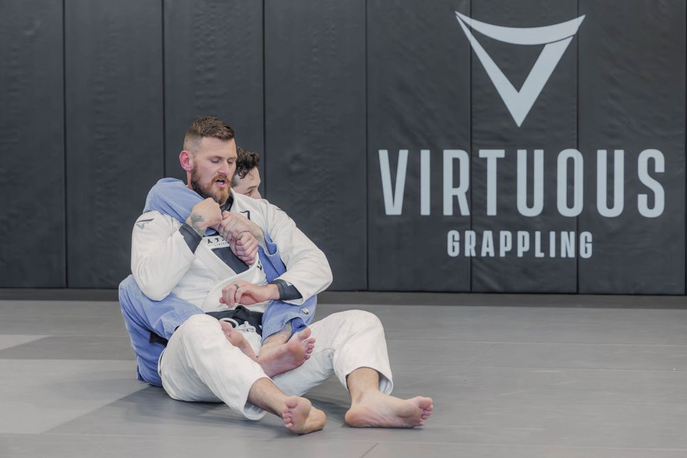 Athletes practicing grappling techniques on mats at Virtuous training facility with logo on wall in background.