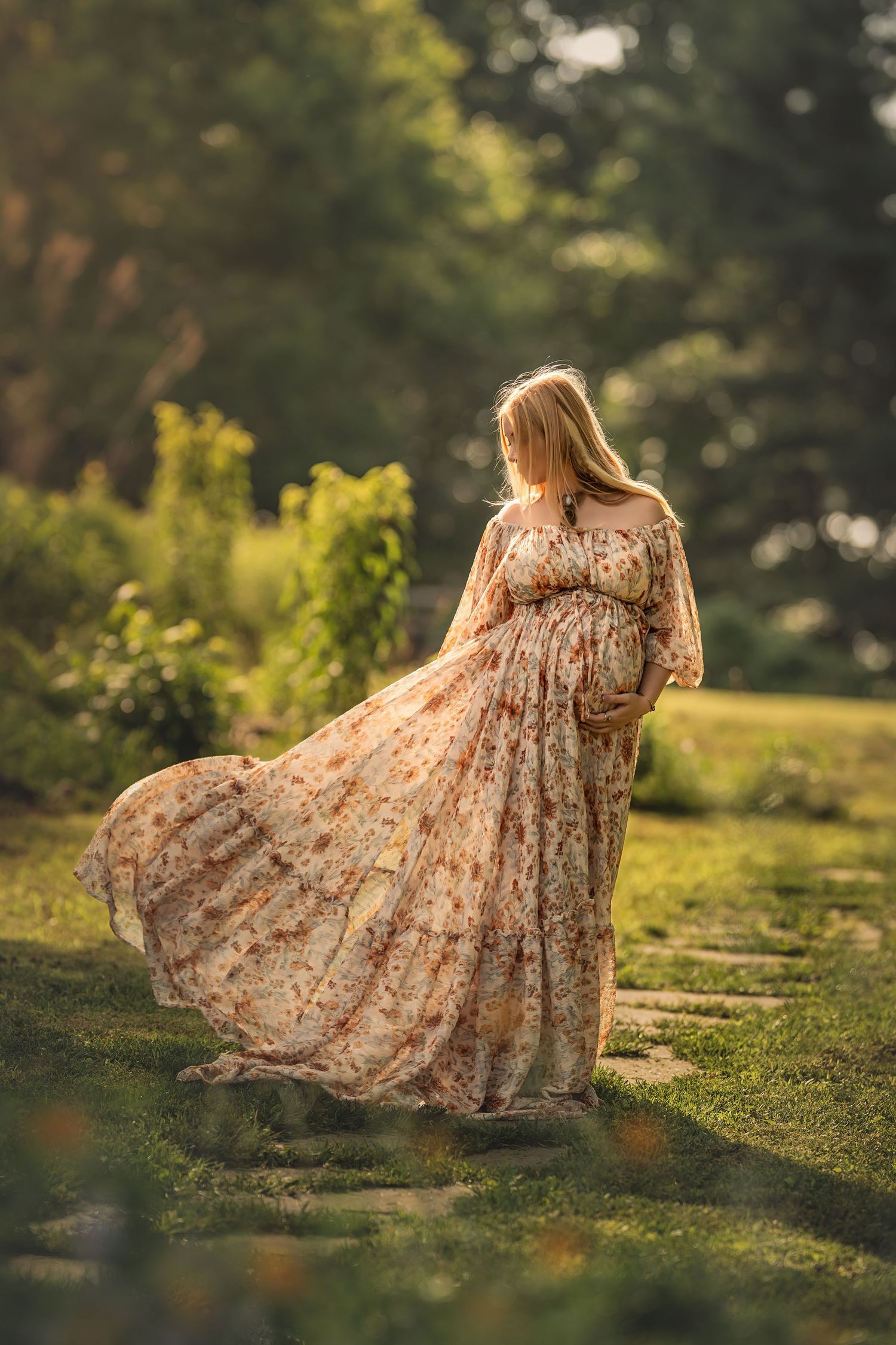 A flowing floral maternity dress catches the sunlight as a woman stands in a dreamy garden setting at golden hour.