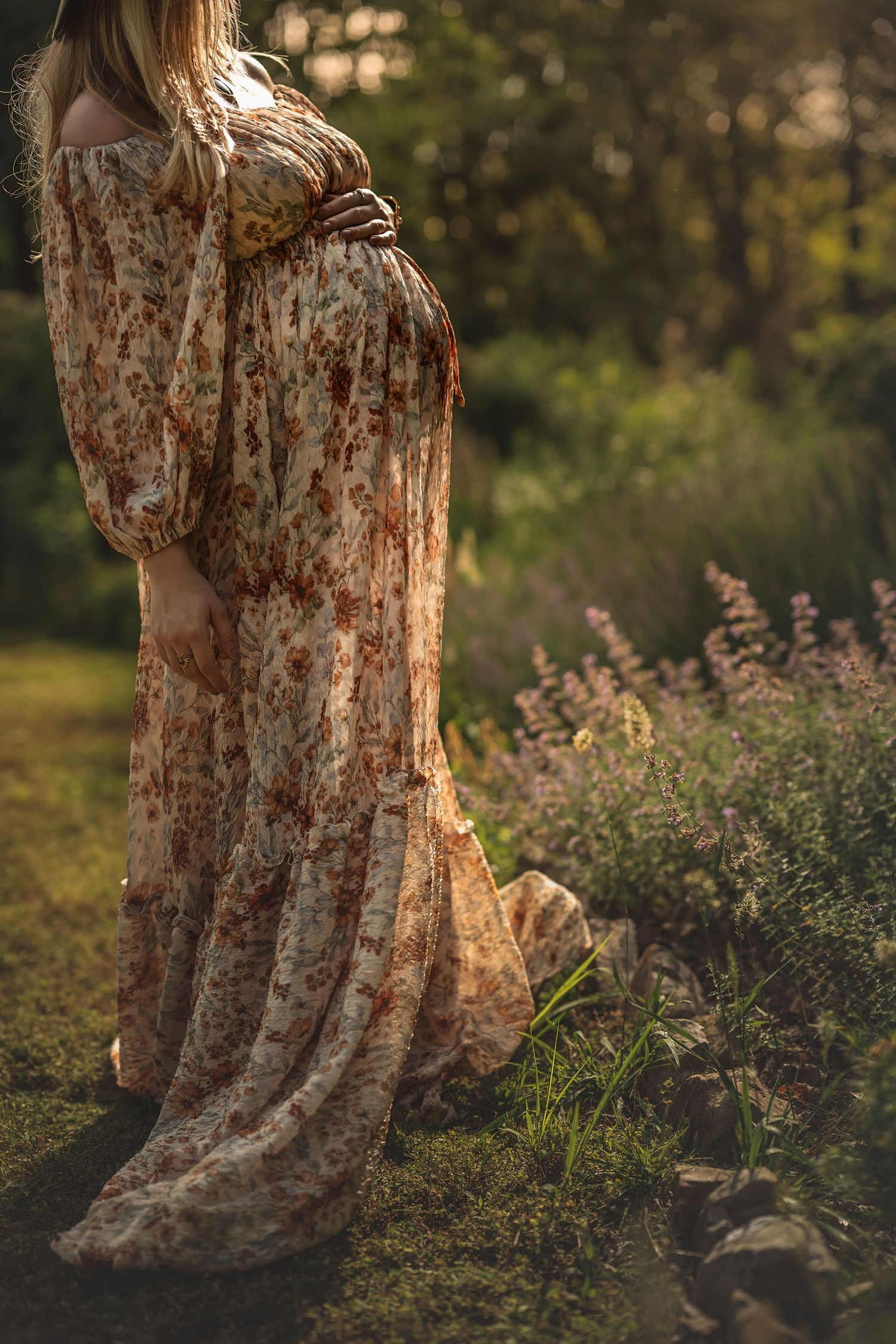 A pregnant woman wearing a flowy floral maxi dress stands backlit in golden sunlight near blooming wildflowers in a garden setting.
