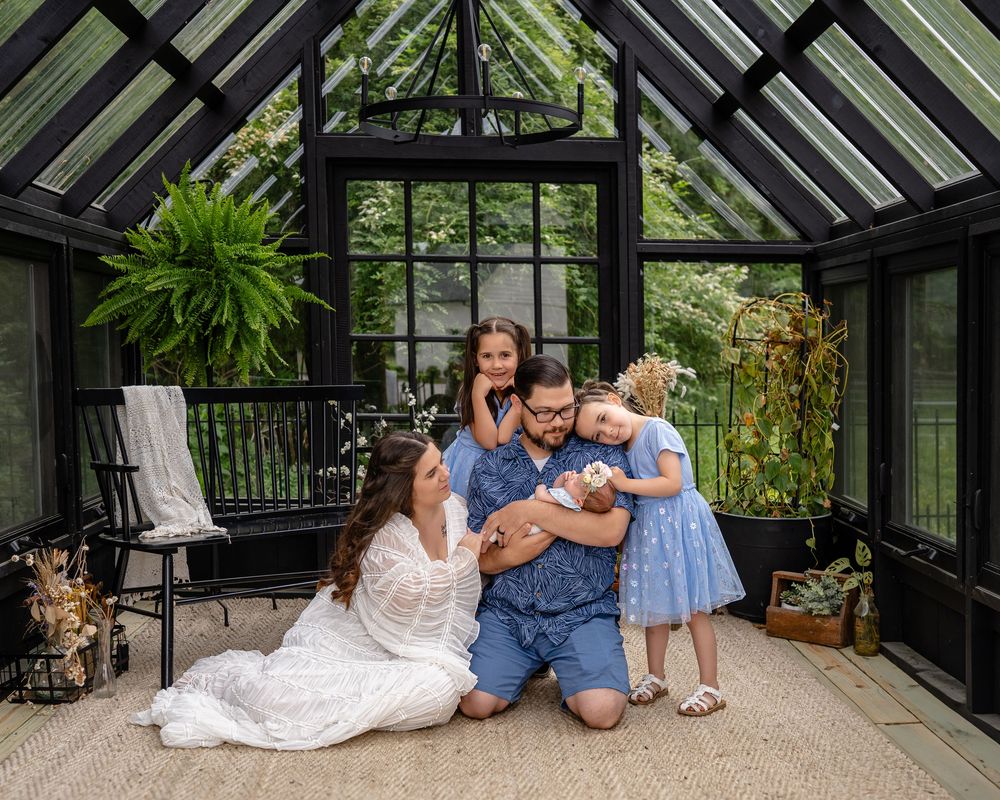 A family shares a joyful moment together inside a sunlit greenhouse with lush greenery and a glass ceiling.