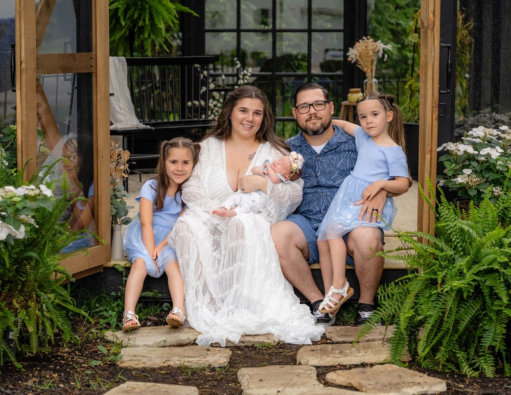 A family sits together on wooden porch steps surrounded by lush green ferns and garden landscaping.