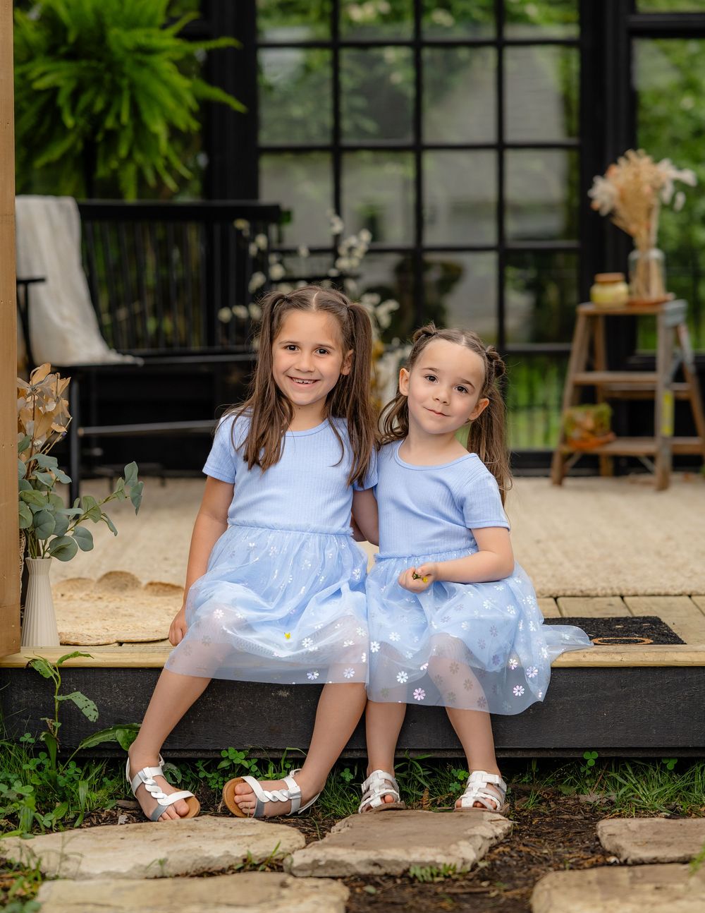 Two young children sitting on rustic wooden steps in coordinated light blue outfits near lush garden landscaping.
