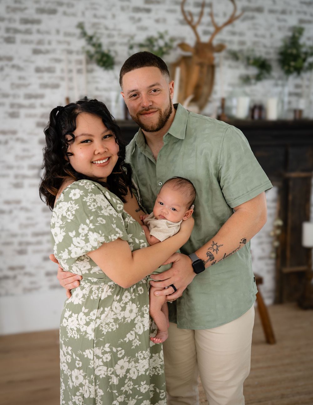 A family of three stands together in front of a white brick wall with rustic decor including a mounted deer head.