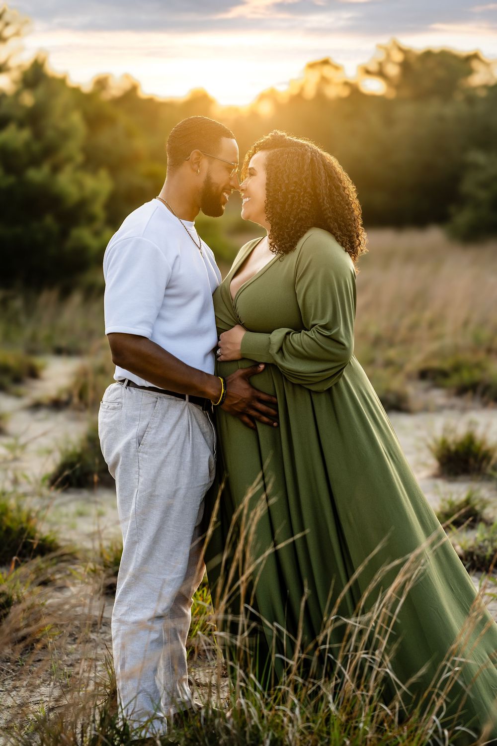 A romantic couple stands in tall grass during golden hour sunset with soft glowing lighting and warm natural tones.