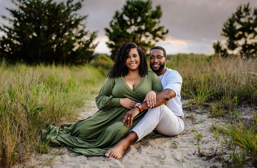 A couple in formal attire sits in a grassy field during golden hour while capturing scenic outdoor portraits.