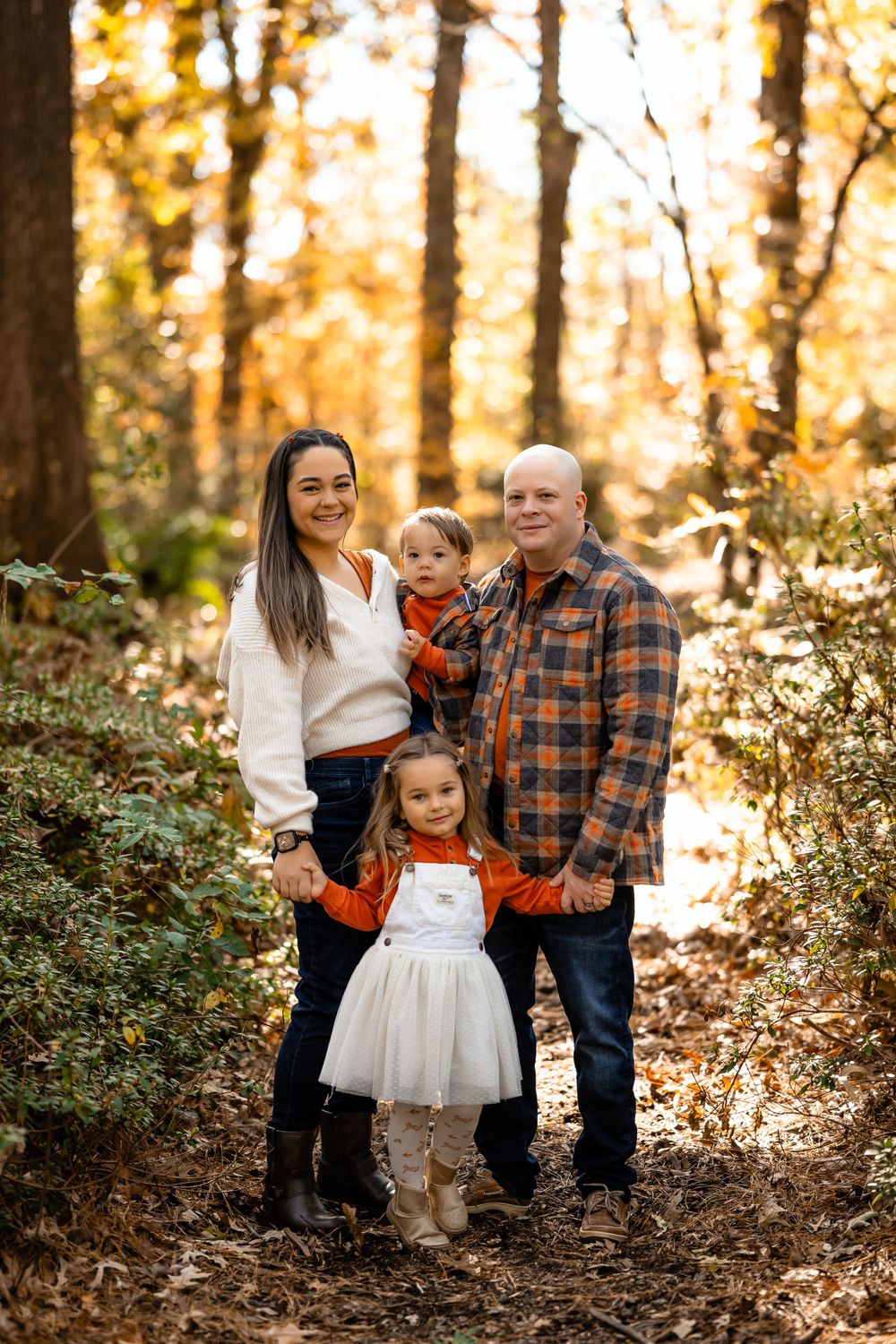 A family in coordinated fall outfits poses together on a leaf-covered forest path.