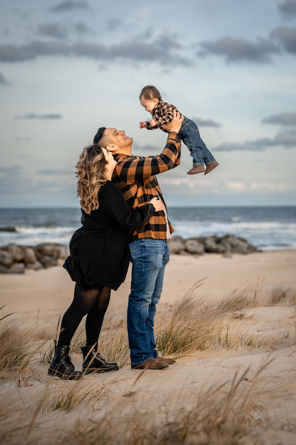 Couple lifts child playfully at beach during sunset with waves crashing in background.