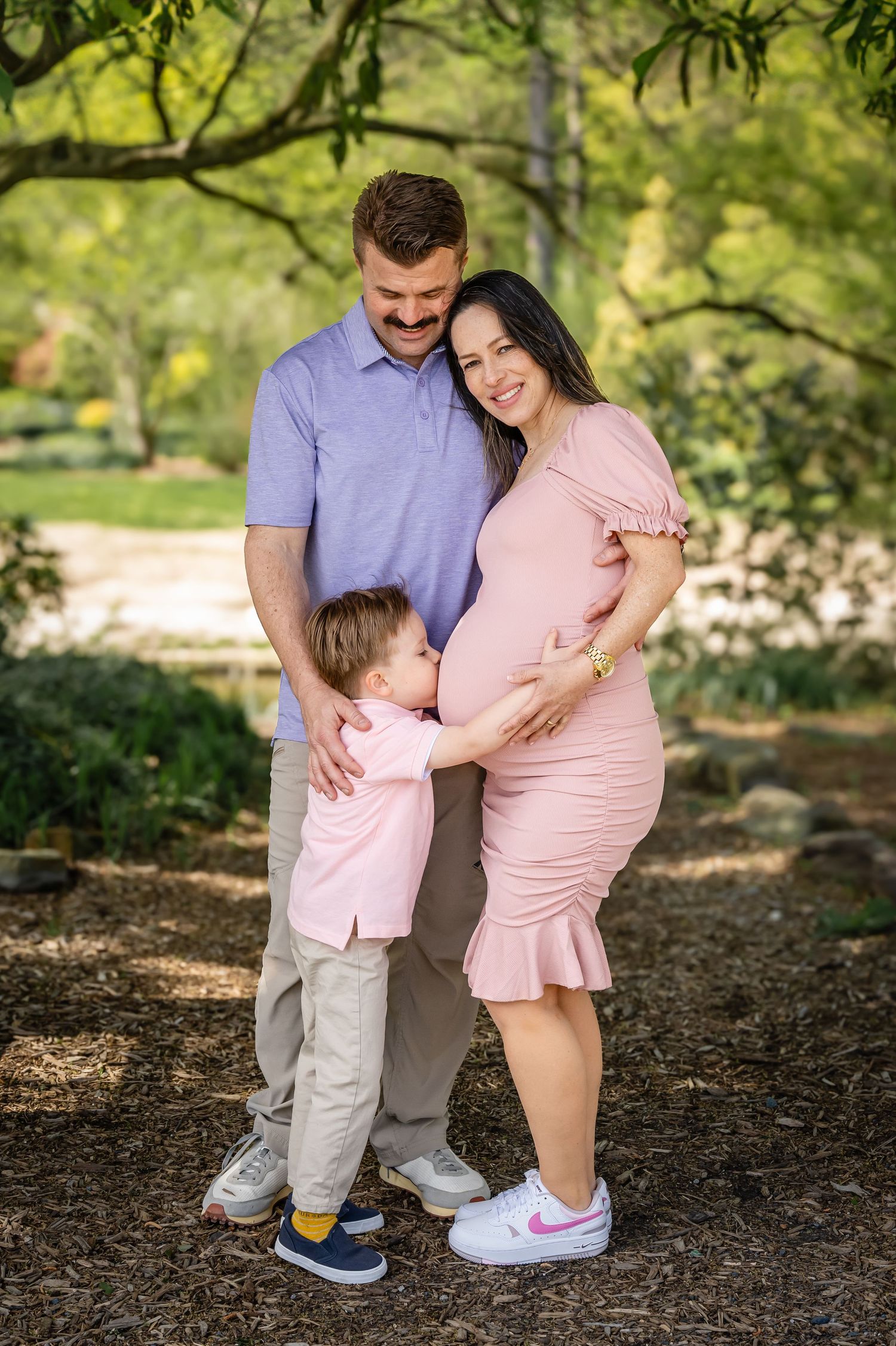 Expecting family in coordinating pink outfits poses together under green trees.