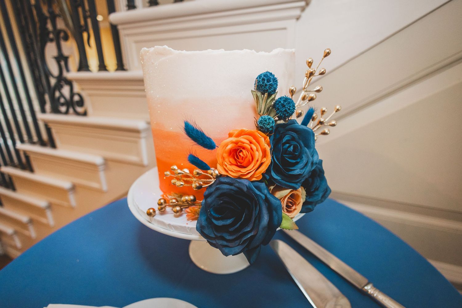 Wedding cake decorated with blue and orange roses sits elegantly on blue tablecloth.