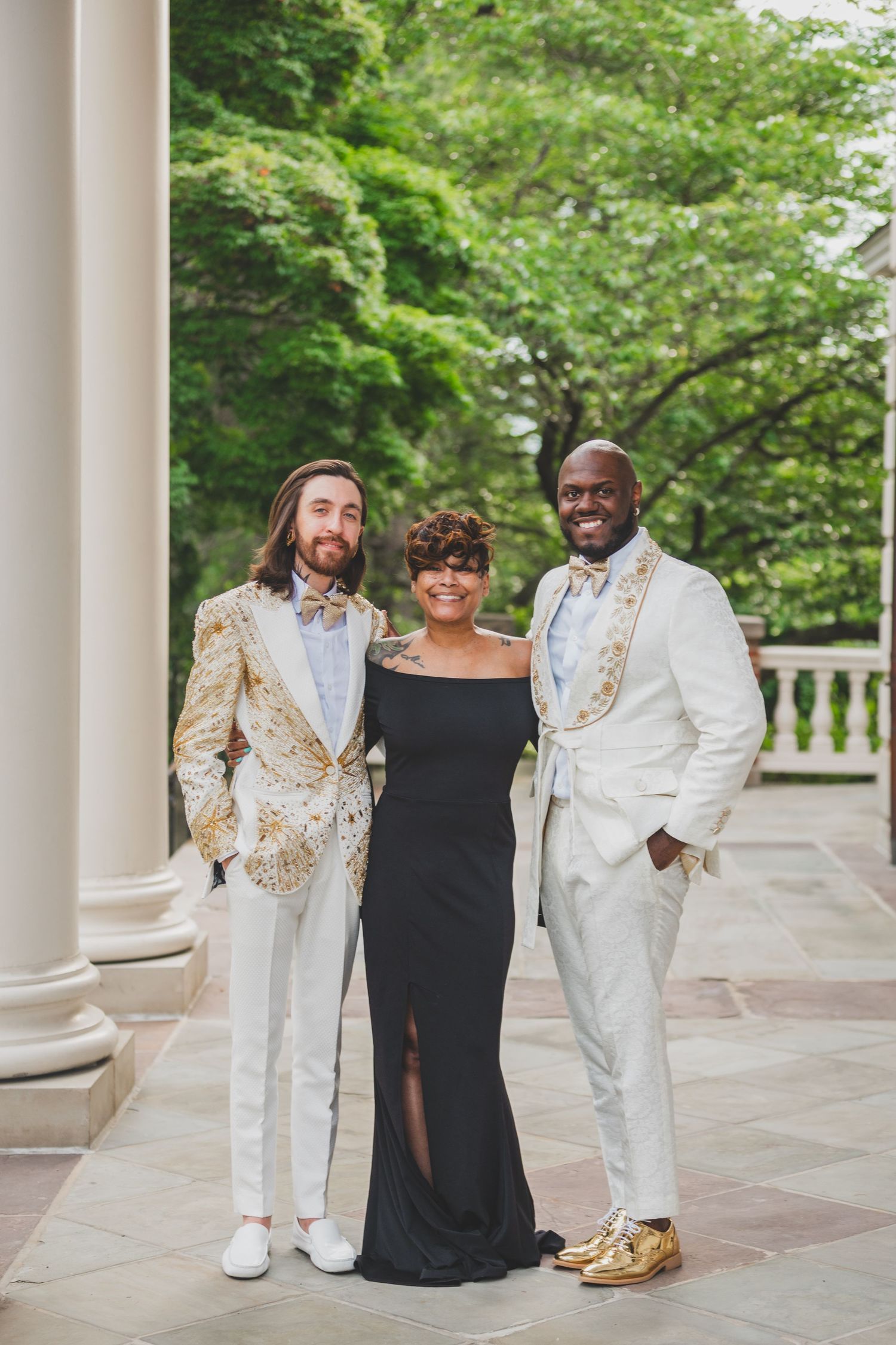 Three people in elegant attire pose together on a columned patio surrounded by lush greenery.