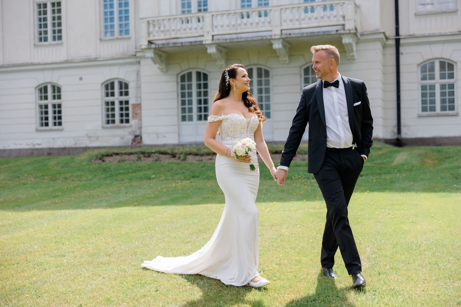 A newlywed couple walks hand in hand across a mansion lawn, with the bride in white and groom in classic tuxedo.