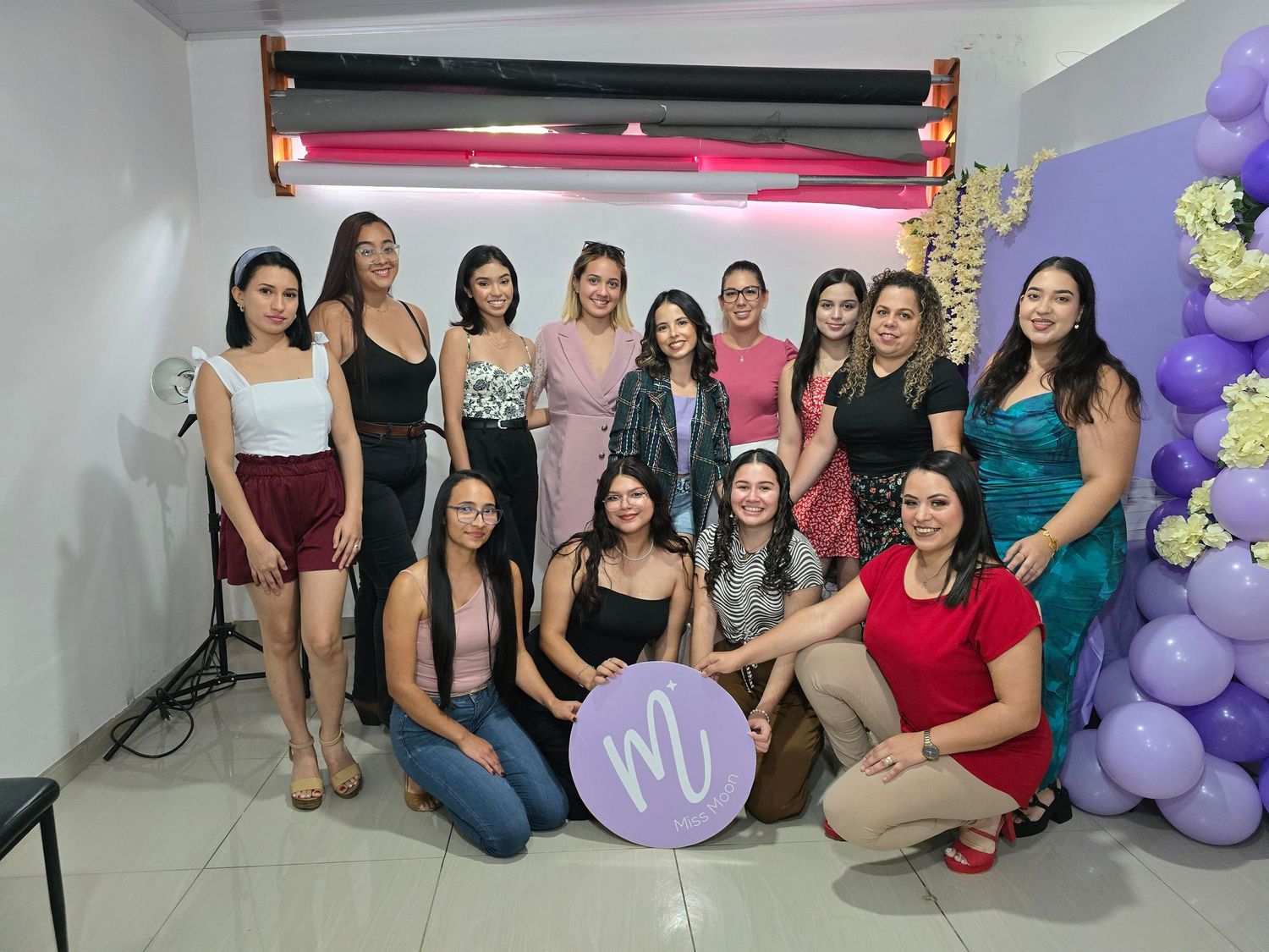 Group photo of women gathered around a purple logo sign at what appears to be a social event or celebration.