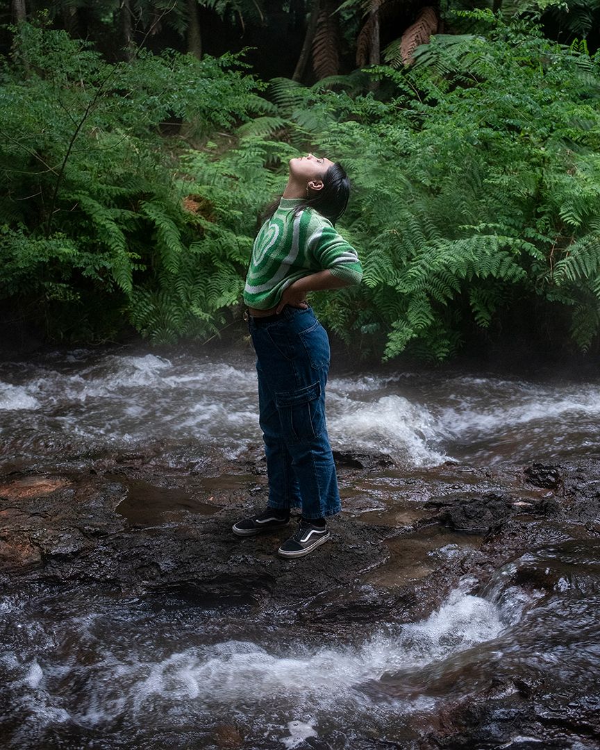 Photograph of a young person staring up to the sky, hands on hips, in the middle of a stream. Ferns and forest