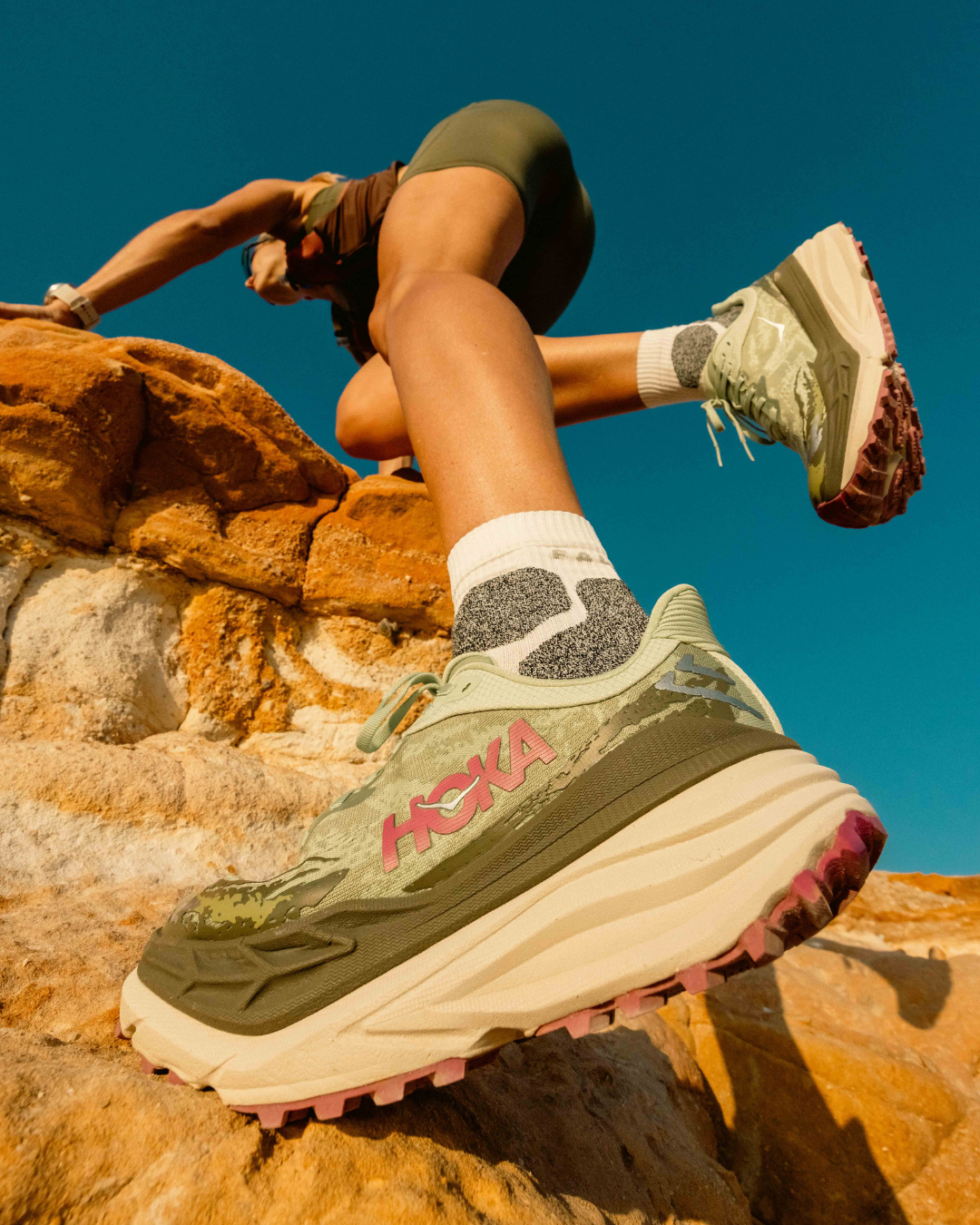 Close-up of a Hoka running shoe on orange rocks against bright blue sky showcasing athletic footwear.