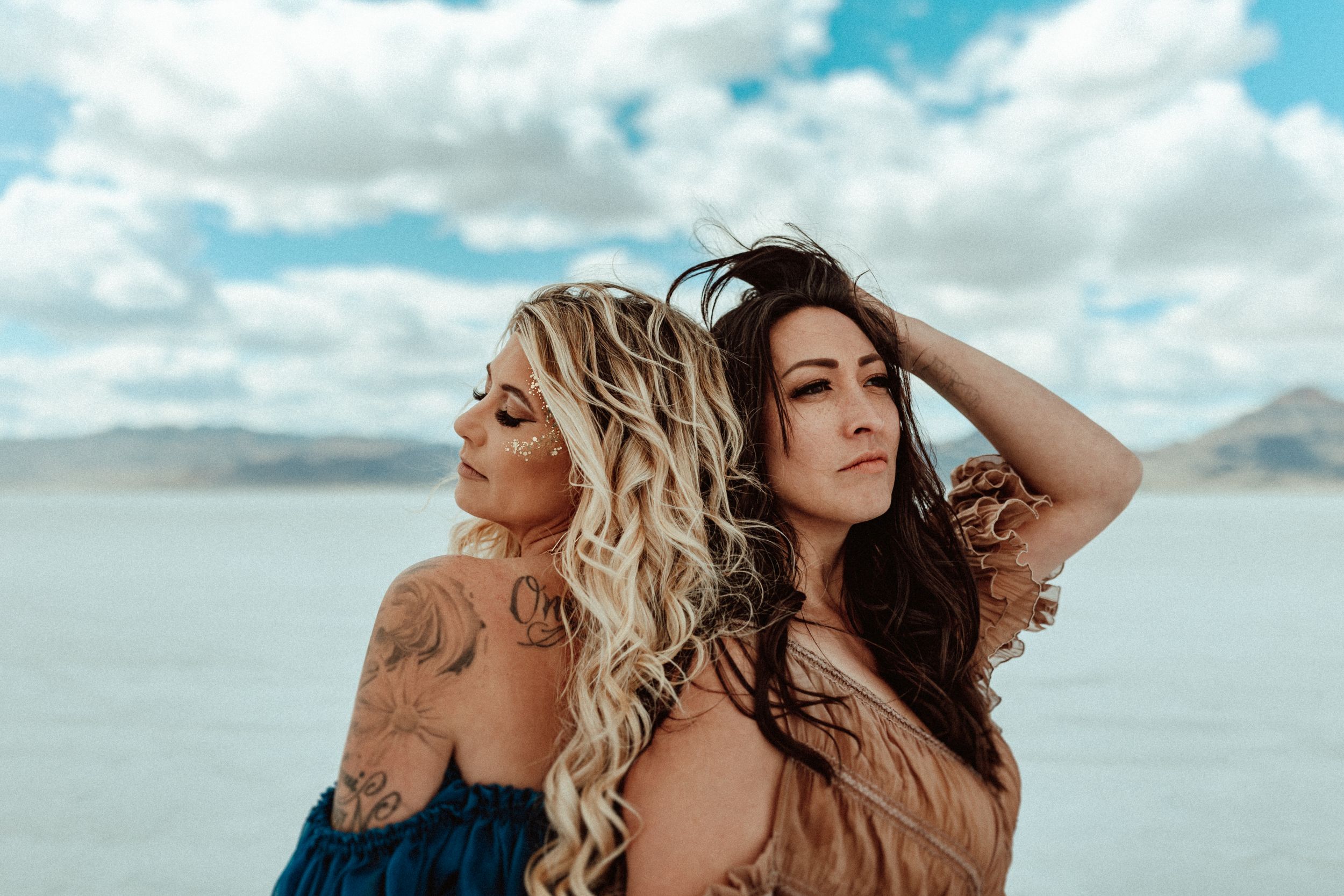 Two women pose against a serene lake backdrop with mountains and cloudy sky.