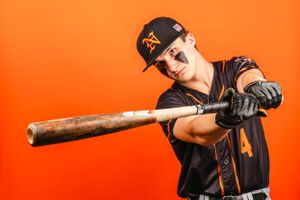 Baseball player in black uniform posing with bat against orange background. Normandy High School Baseball.