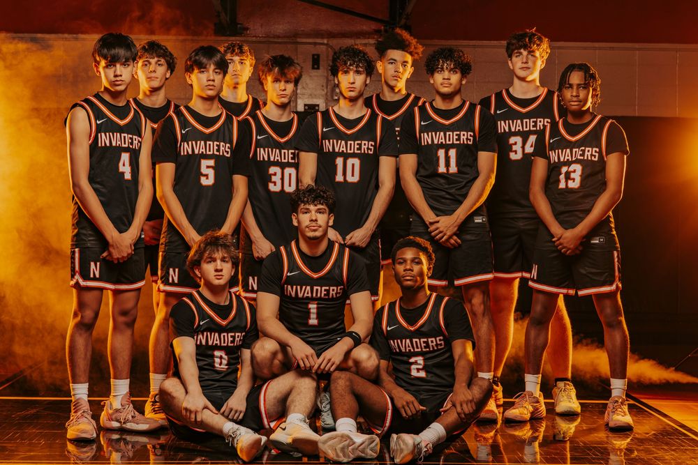 A basketball team poses together in dark uniforms against a warm-toned background in a gym setting. Normandy High School Basketball Team.