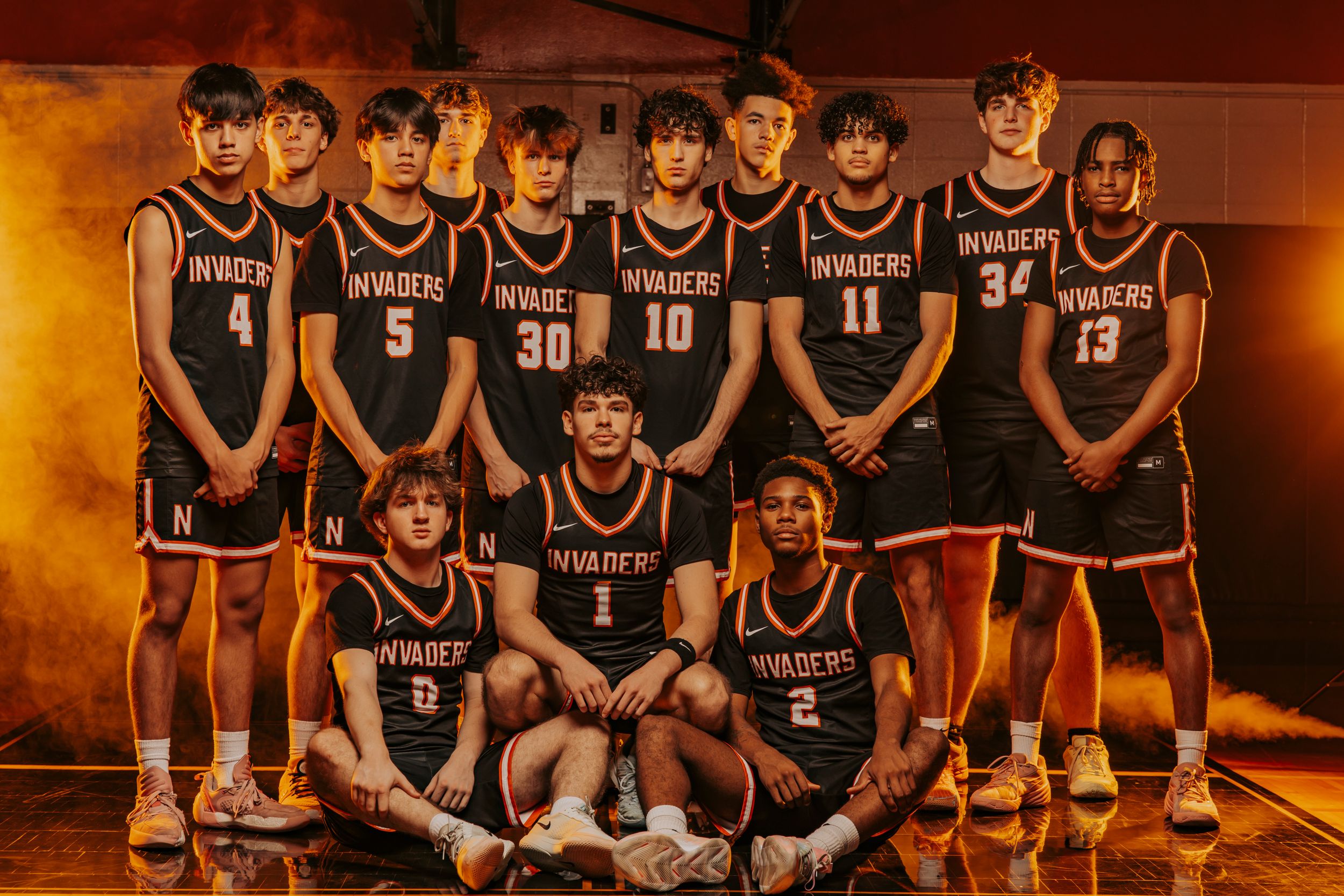 A basketball team poses together in dark uniforms against a warm-toned background in a gym setting. Normandy High School Basketball Team.