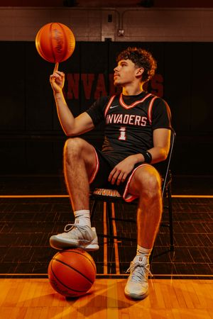 Basketball player in black jersey spins ball on finger while sitting on wooden court in dramatic lighting. Normandy High School Basketball